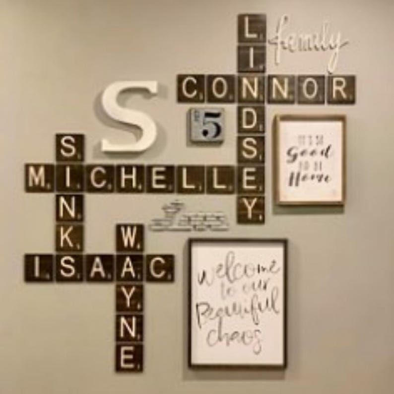 May include: A wall display featuring dark brown wooden tiles with white letters arranged like a crossword puzzle, spelling names including Michelle, Isaac, Wayne, Lindsey, and Connor. It includes a large white letter S, a calendar tile, and two framed signs.