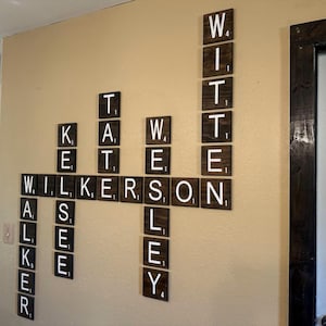 May include: A wall-mounted display of dark brown wooden square tiles arranged in a crossword puzzle format. Each tile features a white letter and a small numerical value in the corner, spelling out the names Walker, Kelsee, Tate, Wesley, and Witten.