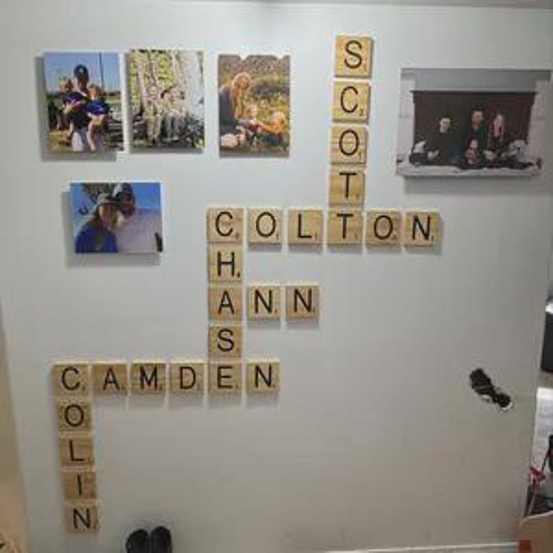 May include: A wall display featuring four rectangular photo canvases and multiple wooden letter tiles arranged in a crossword pattern. The tiles spell out the names Scott, Colton, Chase, Ann, Camden, and Colin. The photos depict various family scenes.
