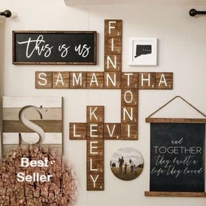 May include: A wall display featuring a rectangular sign with the text this is us, a crossword-style arrangement of wooden tiles spelling names, a square framed map, a circular photo, and a hanging sign with the text and together they built a life they loved.