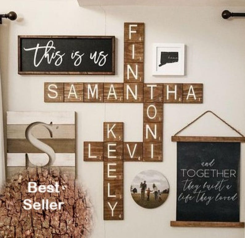 May include: A wall display featuring a rectangular sign with the text this is us, a crossword-style arrangement of wooden tiles spelling names, a square framed map, a circular photo, and a hanging sign with the text and together they built a life they loved.