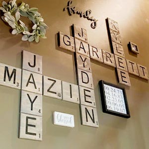 May include: Wooden wall art featuring personalized names and dates in a crossword puzzle design. The names "Jayden" and "Garrett" are spelled out with individual letter tiles. A small framed display includes dates. A decorative wreath and the word "family" are also present.