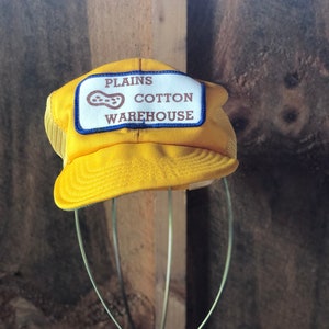May include: A yellow trucker hat with a mesh back and a curved brim. The hat features a rectangular patch with the words "PLAINS COTTON WAREHOUSE" and a peanut graphic. The hat is displayed on a wire stand against a wooden background.