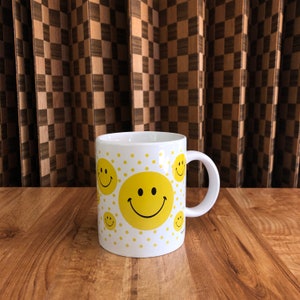May include: White ceramic mug featuring yellow smiley faces and yellow polka dots. The mug has a white handle and is set against a wooden surface and a brown and tan checkered background. A cheerful and fun design.
