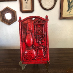 May include: A vintage red wall plaque featuring a relief design of a kitchen scene. The plaque includes a lamp, clock, and teapot. The plaque is displayed on a brass easel, showcasing a rustic, country aesthetic.