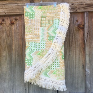 May include: A round, patchwork-style textile with a cream-colored fringe. The fabric features a mix of geometric and floral patterns in shades of green and tan. The textile is hanging on a wooden background.