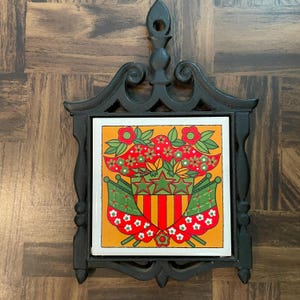 May include: A square ceramic tile trivet set in a black cast iron frame with a hanging loop. The tile features a yellow background with a central shield design containing three green stars and vertical red and yellow stripes, flanked by green flags.