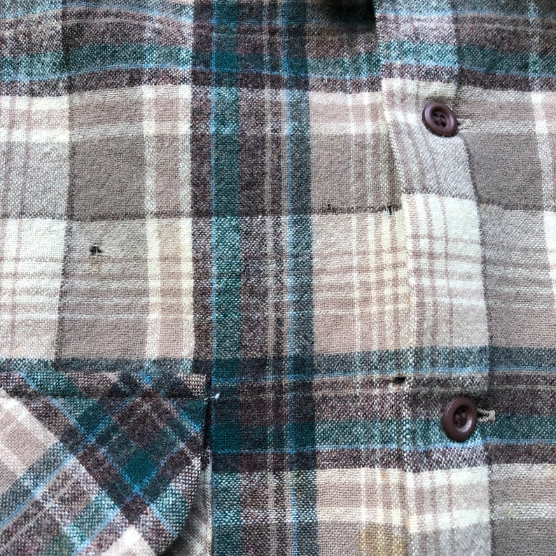May include: Close-up of a button-down shirt with a plaid pattern in shades of gray, teal, and beige. The shirt features two brown buttons and a pocket detail. The fabric appears to be a woven material.