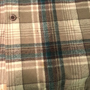 May include: Close-up of a plaid fabric in shades of beige, gray, teal, and brown. The pattern is a classic grid design with varying widths of lines. A single, round, brown button is visible on the fabric.