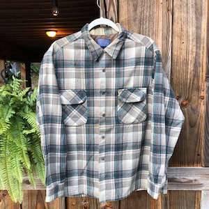 May include: A plaid button-up shirt in shades of beige, teal, and brown, with two chest pockets. The shirt is hanging on a white hanger against a wooden background. The shirt has a collar and buttoned cuffs.
