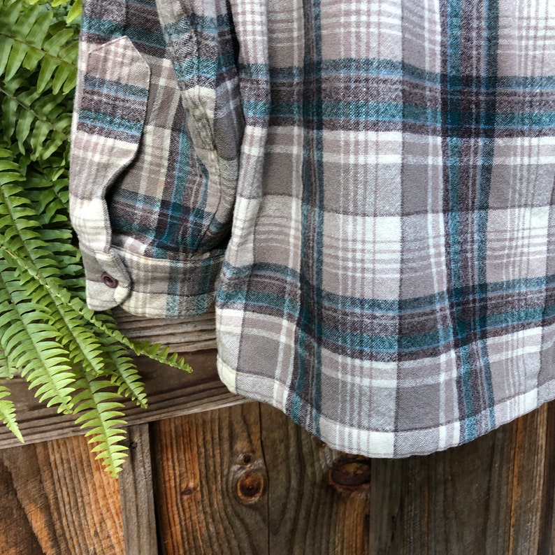 May include: Close-up of a gray, teal, and white plaid flannel shirt. The shirt has a buttoned cuff and a pocket. The fabric is soft and appears to be made of a warm material. The shirt is displayed on a wooden surface.