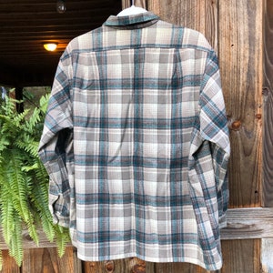 May include: A long-sleeved flannel shirt with a plaid pattern in shades of gray, teal, and white. The shirt is hanging on a white hanger, showcasing the back of the garment. The fabric appears soft and comfortable.