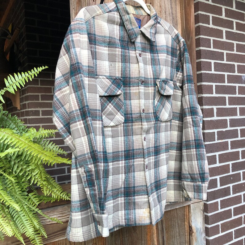 May include: A button-down plaid shirt in shades of gray, teal, and cream, with two chest pockets. The long-sleeved shirt is hanging on a wooden door. The shirt has a classic collar and buttoned cuffs.