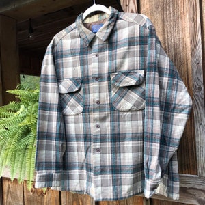 May include: A button-down plaid shirt in shades of gray, teal, and white. The shirt has two chest pockets and a collared neckline. The fabric appears to be a soft, woven material. The shirt is hanging on a white hanger.