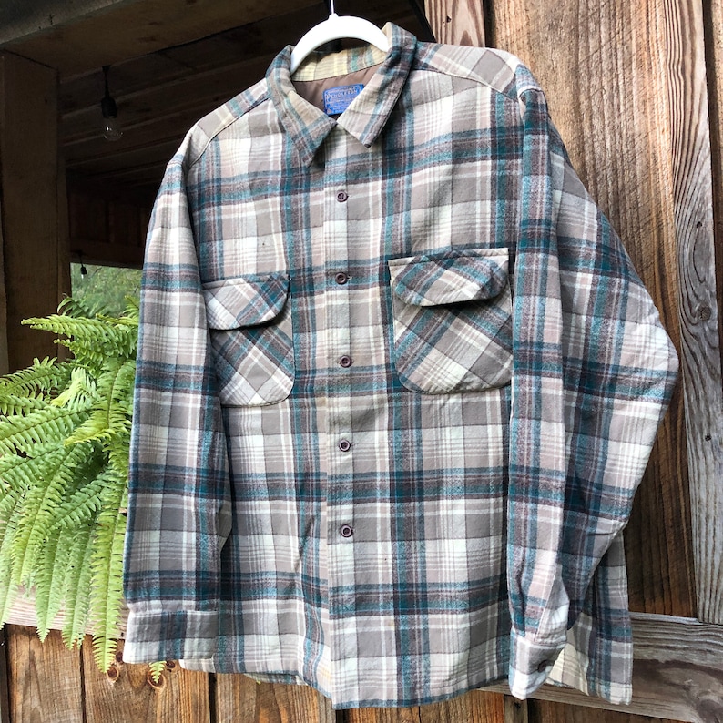 May include: A button-down plaid shirt in shades of gray, teal, and white. The shirt has two chest pockets and a collared neckline. The fabric appears to be a soft, woven material. The shirt is hanging on a white hanger.