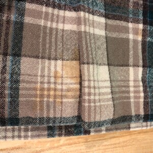 May include: Close-up of a plaid fabric in shades of gray, teal, and white. The pattern is a classic check design, with the fabric appearing to be a woven material. The texture of the fabric is visible, and the overall look is casual and comfortable.