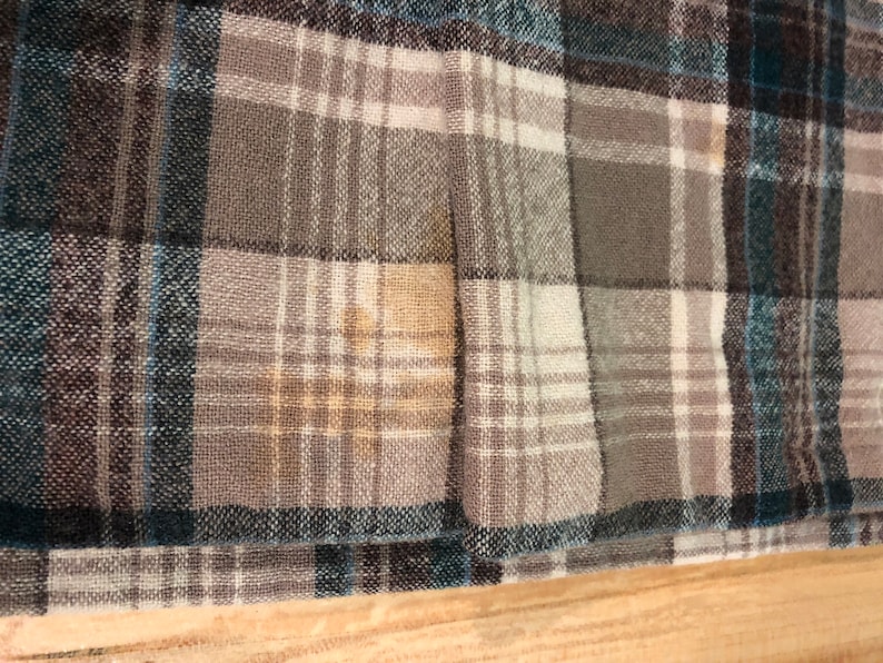 May include: Close-up of a plaid fabric in shades of gray, teal, and white. The pattern is a classic check design, with the fabric appearing to be a woven material. The texture of the fabric is visible, and the overall look is casual and comfortable.