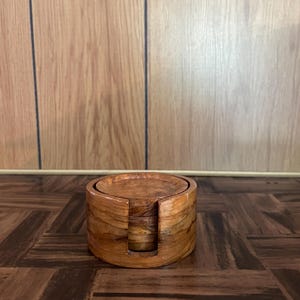 May include: A cylindrical wooden holder containing a stack of circular wooden coasters. The holder features a vertical cutout slot on the front side, and both the holder and the coasters exhibit a natural light brown wood grain pattern.