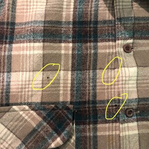 May include: Close-up of a button-down flannel shirt with a plaid pattern in shades of brown, teal, and cream. The shirt has a chest pocket and brown buttons. The fabric appears to be a soft, woven material.