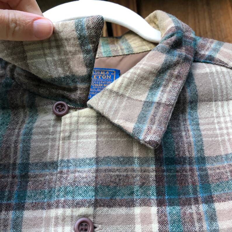 May include: Close-up of a plaid button-up shirt in shades of brown, teal, and beige. The shirt has a collar and two visible buttons. A blue tag with white text is visible. The shirt is hanging on a white hanger.
