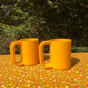 May include: Two bright yellow mugs with large handles sit on a floral patterned tablecloth. The mugs are made of a solid, opaque material and have a simple, cylindrical shape. The background features green foliage.