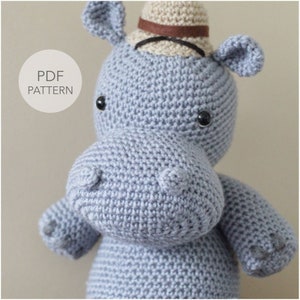 May include: A crocheted gray hippopotamus wearing a brown and beige hat. The hippopotamus has black eyes and a small, rounded nose.