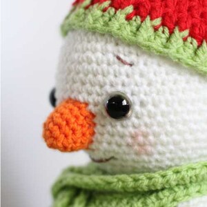 May include: Close-up of a crocheted snowman with a red and green hat, an orange nose, and black eyes. The snowman is wearing a green scarf.