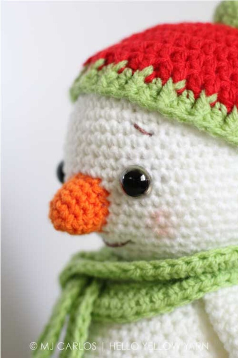 May include: Close-up of a crocheted snowman with a red and green hat, an orange nose, and black eyes. The snowman is wearing a green scarf.