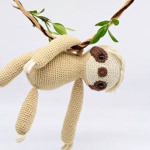 May include: A crocheted sloth toy hanging from a brown and beige rope with green leaves. The sloth is beige with brown eyes and a white face.