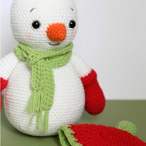 May include: A white crocheted snowman with a green scarf and red mittens. The snowman is wearing a red and green crocheted hat.