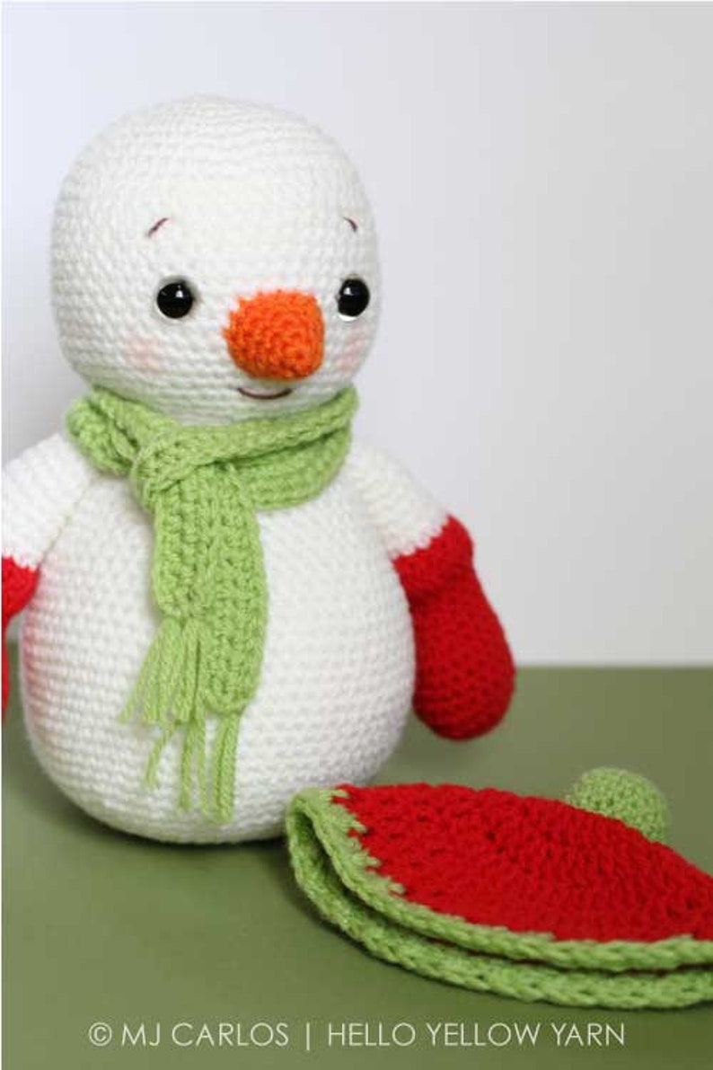 May include: A white crocheted snowman with a green scarf and red mittens. The snowman is wearing a red and green crocheted hat.
