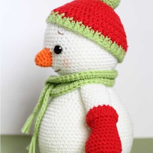 May include: A crocheted snowman wearing a red and green hat with a white pom pom, a green scarf, and a red mitten.