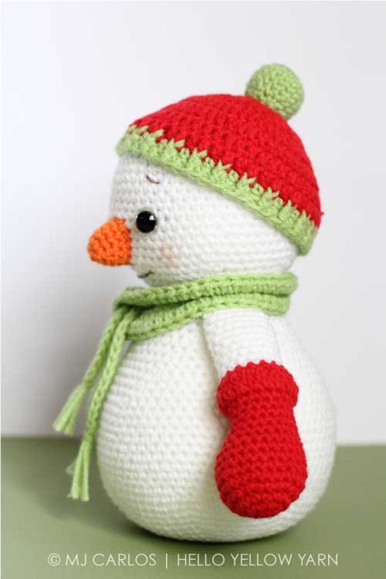 May include: A crocheted snowman wearing a red and green hat with a white pom pom, a green scarf, and a red mitten.