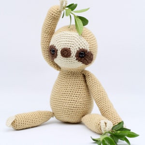 May include: A crocheted sloth toy with brown eyes and a beige body. The sloth is holding a green leafy branch in its hand.