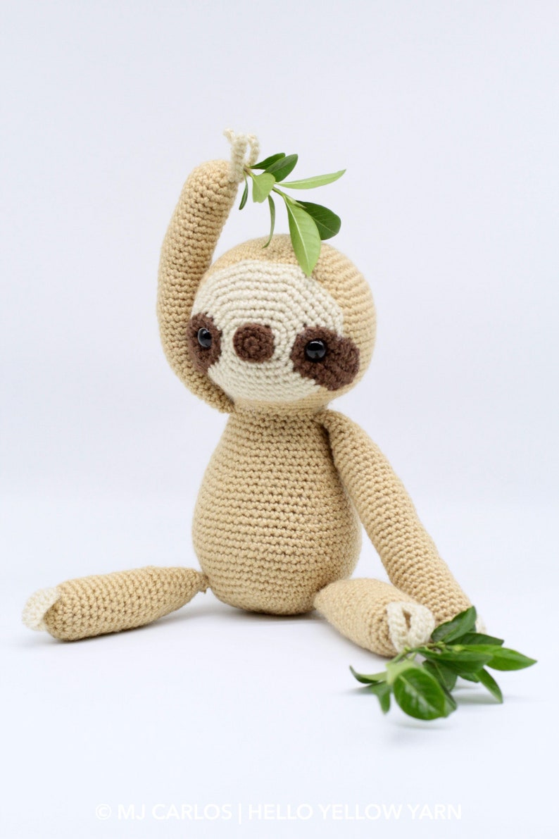 May include: A crocheted sloth toy with brown eyes and a beige body. The sloth is holding a green leafy branch in its hand.