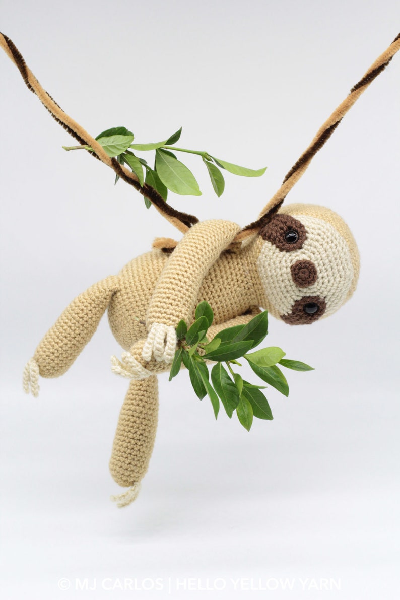 May include: A crocheted sloth toy hanging from a brown and tan branch with green leaves. The sloth is beige with brown eyes and a white belly.