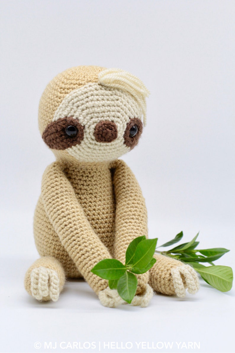 May include: A crocheted sloth toy with brown eyes, a beige body, and white feet. The sloth is sitting on a white background with green leaves.