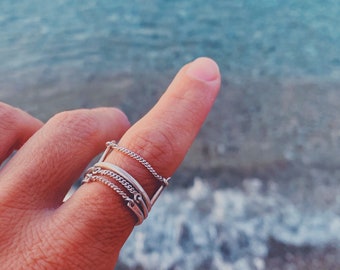 Könnte beinhalten: Ein silberner Ring mit mehreren übereinander gestapelten Kettengliedern. Der Ring wird an einem Finger getragen, der auf einen verschwommenen Hintergrund aus blauem Wasser zeigt.