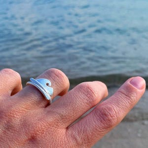 Può includere: Un anello d'argento con un design a onda. L'anello è indossato su un dito e lo sfondo è un'immagine sfocata di acqua.