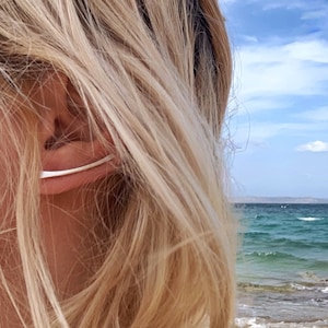 Könnte beinhalten: Nahaufnahme eines Frauenohrs mit einem silbernen Ohrring. Der Ohrring ist ein dünnes, gebogenes Stück Metall, das auf dem oberen Teil des Ohrs sitzt. Die Haare der Frau sind blond und fließend.