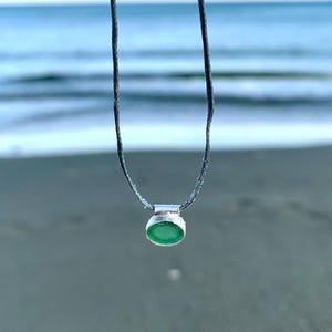 May include: A silver pendant necklace with a small, oval, green gemstone. The pendant is suspended from a thin, dark gray cord. The background is a blurred view of the ocean and beach.