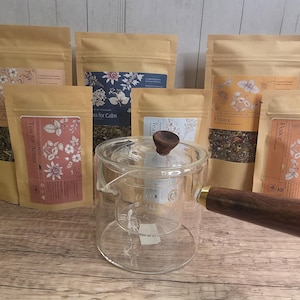 May include: A clear glass teapot with a wooden handle and lid is surrounded by several brown paper bags of tea. The bags have labels with floral designs and text, including "Tea for Active" and "Peace". The tea inside the bags is visible.