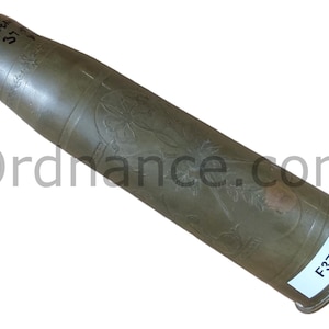 May include: A vintage brass artillery shell casing with etched floral designs. The shell has a cylindrical shape with a slightly tapered end. The text "FREA 37" is visible on the top. A white label with "F37" is on the bottom.