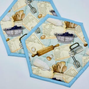 May include: Two hexagonal quilted pot holders with a light blue border. The fabric features a vintage kitchen theme with illustrations of recipes, eggs, a rolling pin, and a hand mixer. A colander of berries and wooden spoons are also depicted.