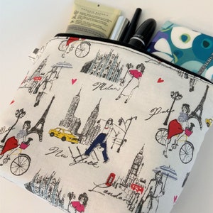 May include: White zippered pouch with a black zipper. The pouch has a white background with a black and gray city scene pattern. The pattern includes the words "Milan", "New York", and "London".