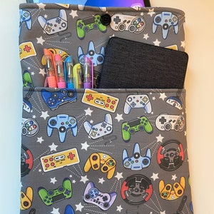 May include: Gray tablet sleeve with a pattern of colorful video game controllers and white stars. The sleeve has a pocket with pens and a small black device. A tablet is visible inside the sleeve.