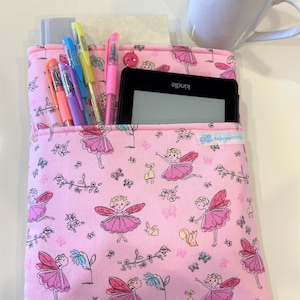 May include: Pink fabric book sleeve with a floral pattern featuring fairies, butterflies, and small animals. The sleeve has a pocket for pens and a larger pocket for an e-reader. The e-reader has the word "Kindle" printed on it.