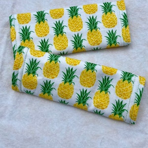 May include: Two white fabric pot holders with a pineapple pattern. The pineapples are yellow with green leaves.