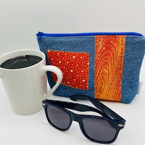 May include: A blue denim zippered pouch with a red and orange patterned fabric insert. The pouch is sitting on a white surface next to a white coffee mug and black sunglasses.