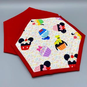 May include: Two red and white fabric pot holders with a hexagon shape. The pot holders feature a white background with a pattern of colorful cupcakes and sprinkles. The cupcakes have Mickey Mouse ears and are decorated with different characters, including Mickey Mouse, Minnie Mouse, and Donald Duck. The pot holders are trimmed with a red fabric border.